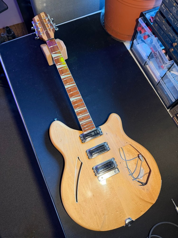 A top-down view of a wooden electric guitar with a natural finish, featuring three pickups and a unique body shape, resting on a black surface with various tools and objects nearby.